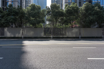 Urban road with asphalt surface, tree-lined fence, and modern high-rise buildings in background © zhu difeng