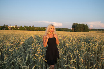 Fototapeta premium Female model with curly blonde hair wearing a black dress stands in a field of wheat during sunset, with soft clouds and a colorful sky in the background