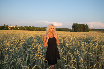 Fototapeta premium Female model with curly blonde hair wearing a black dress stands in a field of wheat during sunset, with soft clouds and a colorful sky in the background