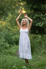 Obraz premium Young woman with long curly hair wearing a white crop top and skirt stands on a forest path, adorned with a floral crown, surrounded by greenery and sunlight