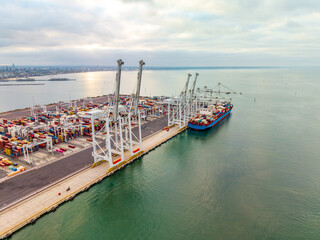 Naklejka premium Aerial view of a busy container ship docked at the Port of Melbourne, with gantry cranes actively handling stacks of colorful shipping containers.