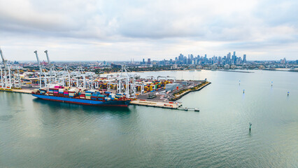 Fototapeta premium An expansive aerial shot captures the busy Port of Melbourne, showcasing a large cargo vessel, numerous cranes, and the distant city skyline.