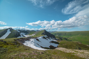 Scenic sunlit view along grassy ridge with white snow cornice to green hill top with rocky sheer crags under lush clouds in blue sky. Lake in mountain cirque in sunny day. High mountains in bright sun © Daniil