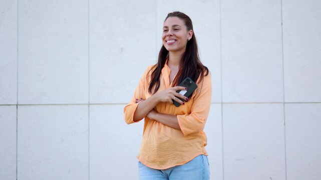Young smiling caucasian woman texting on smartphone and looking away against a white modern wall outdoors in slow motion