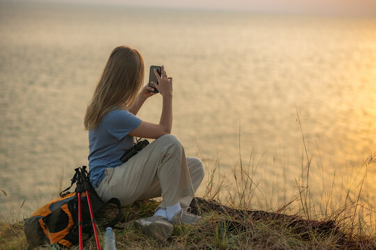 Hiker on grassy hill overlooking sea sunset taking photo with her smartphone backpack and trekking beside representing travel adventure and peaceful outdoor lifestyle.