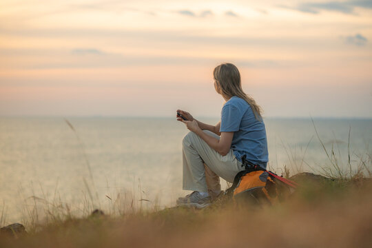 Hiker on grassy hill overlooking sea sunset taking photo with her smartphone backpack and trekking beside representing travel adventure and peaceful outdoor lifestyle.