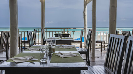Fototapeta premium Seaside cafe. The tables are set. There are cutlery and glasses on the napkins. Wooden chairs. Behind the latticed railings is an aquamarine ocean. Sun umbrellas on the beach. Cuba. Varadero. Resort. 