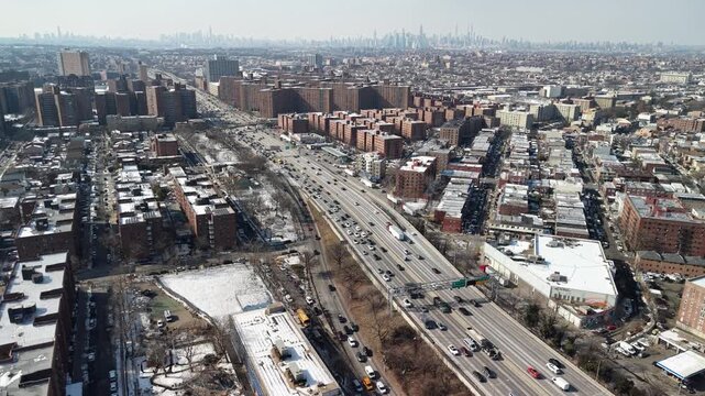 High angle drone footage capturing cars and trucks driving along a busy highway through a snowy new york city borough, with the iconic manhattan skyline visible on the distant horizon