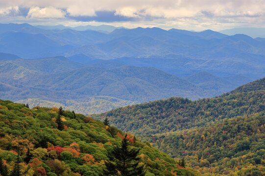 A panoramic view of the Blue Ridge Mountains in North Carolina during autumn, showcasing the vibrant fall foliage with layers of mountains fading into the distance under a cloudy sky.