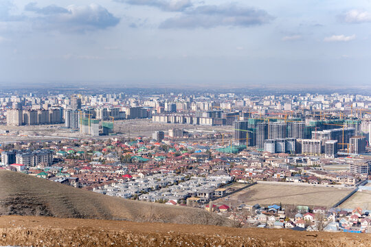 A high-angle, wide shot of a developing city skyline in Bishkek, Kyrgyzstan, where modern high-rise buildings under construction contrast sharply with older, low-rise residential areas in the foregrou