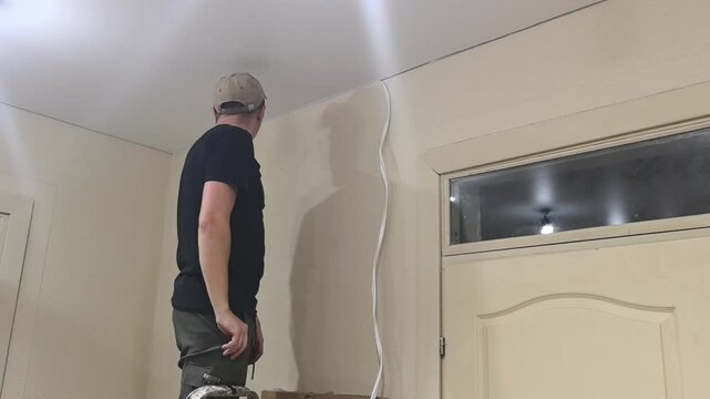 A man is installing decorative baseboards along a suspended ceiling in a room. He uses a tool to attach the baseboards to the wall. The scene takes place in the house in the evening.