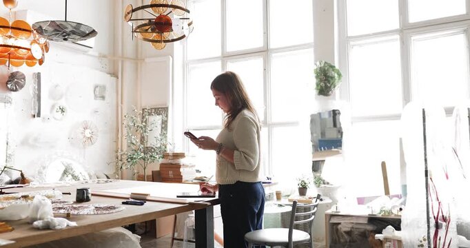 Female artist sketching, working on design using digital tablet and smartphone, standing in sunlit studio at workbench with tools, materials and glass pieces. Modern creative workflow, craftsmanship