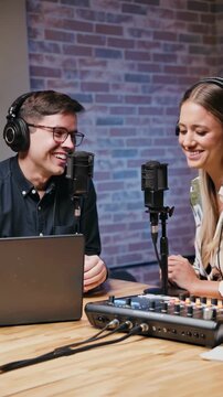 Two people engaged in a lively podcast recording session, smiling and interacting with microphones and headphones in a studio setting, vertical shot