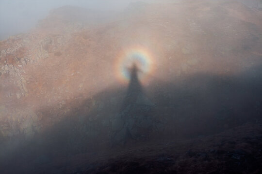 Mysterious silhouette of the optical illusion Brocken Spectre or Brocken bow, mountain ghost surrounded by fog and rainbow light in a serene landscape at dawn.