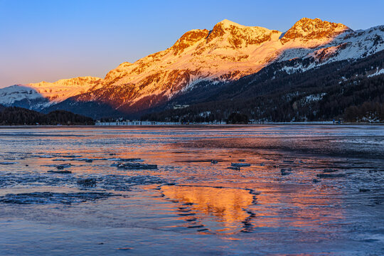 Sunset over the snow-covered mountain with color reflection on the frozen lake ice blocks on the Silsersee Lake in St. Moritz, Grisons, Switzerland
