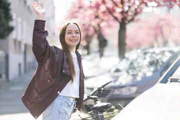 Happy woman greeting neighbors, waving hand on residential city street in spring. Joyful urban lifestyle moment with cherry blossom, blooming trees © Mary Anlens