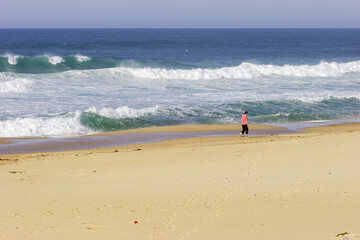 Obraz premium A lone person walks along a sandy beach with waves crashing in the ocean.
