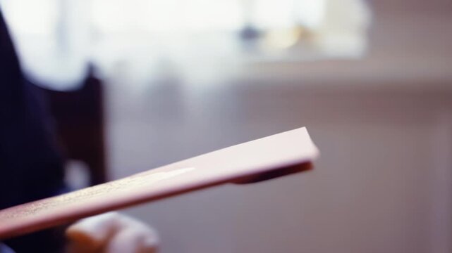 Hand of a person holding a pink envelope with gold embellishments, transitioning to another hand receiving it, showcasing a delicate exchange in a softly lit indoor setting