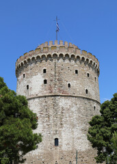 White Tower with Greek Flag between Trees with Blue Sky Background in Thessaloniki, Greece