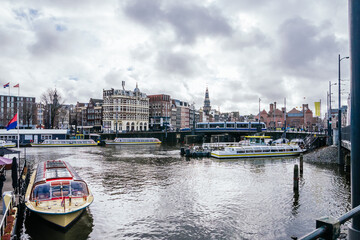 Fototapeta premium Amsterdam canal scene with tour boats cruising past historic waterfront buildings and a tram crossing a bridge under a cloudy sky, capturing urban travel and sightseeing