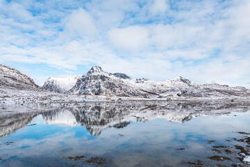 Snow-covered Lofoten peaks reflect perfectly in the calm Arctic water, creating a serene winter landscape with bright sky and distant cabins in the northern Norwegian wilderness © Fauren