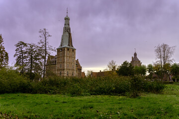 Ein markanter Schlossturm ragt majest&auml;tisch hinter dichten gr&uuml;nen Str&auml;uchern hervor, w&auml;hrend sich der Himmel in ein dramatisches, wolkenverhangenes Violett h&uuml;llt.