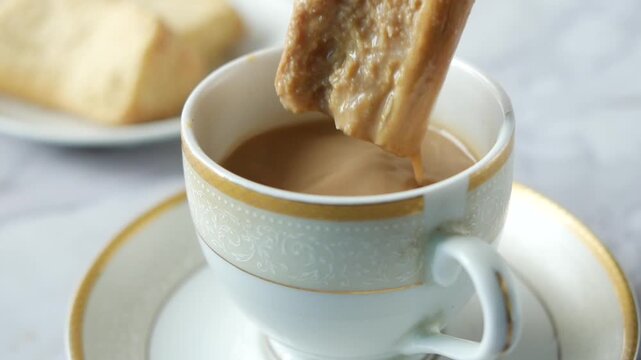 Close up slow motion footage of a hand dipping a biscuit into a cup of tea, with another biscuit resting in the background