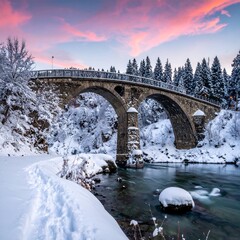 Stone bridge spans a river in winter, snowy banks and evergreens under a pastel dawn