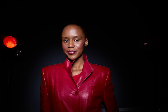 Confident African woman with dark skin wearing a red leather jacket poses in a studio with dramatic lighting. The portrait highlights elegance, fashion, and empowerment concepts.