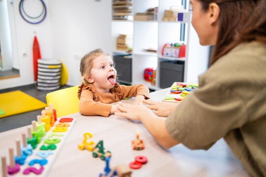 Young child learning articulation exercises with a speech therapist, practicing tongue movements during a developmental therapy session in a bright playroom