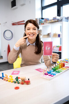 Speech therapist teaching pronunciation with an ee alphabet card, pointing to her mouth while working with a child in a colorful classroom full of educational toys and games