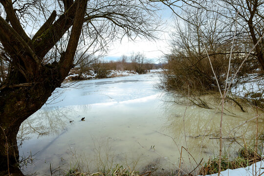 ​A picturesque winter landscape with a frozen river surrounded by bare trees and remnants of snow on the banks under a cloudy sky.