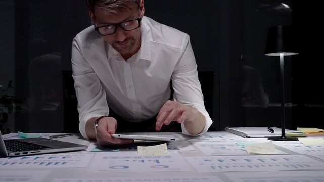 Businessman leans over a desk strewn with charts and reports, working on a tablet in a dim office late at night, reflecting overtime, focus, and relentless dedication