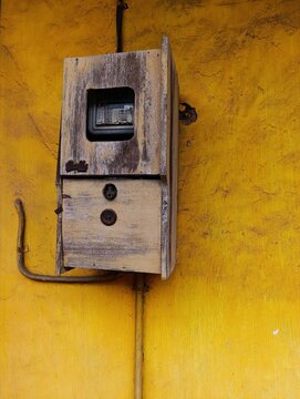 This is an old-fashioned wooden box that covers an electrical energy meter (electrometer) mounted on a wall, worn orange in color and with a rough texture.