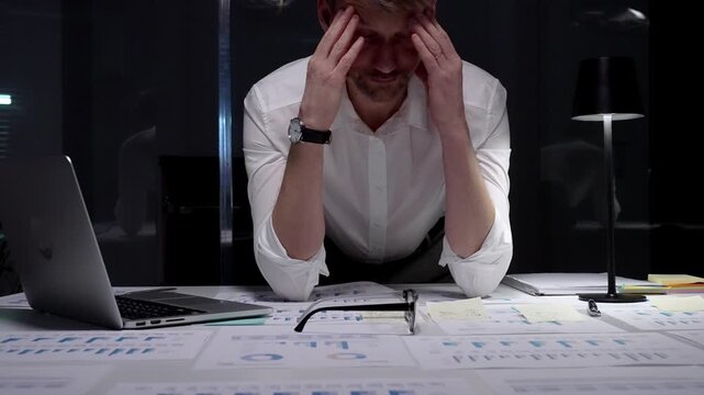 Stressed businessman suffering from overwhelming burnout and work overload, working late into the night, feeling frustrated with deadline pressure in a dark office environment