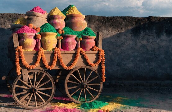 Traditional wooden cart loaded with jute sacks and clay pots of vibrant Holi color powder against a rustic mud wall