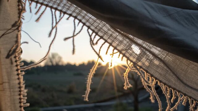 Massive pile of discarded clothing towers in arid landscape concept. Sunset view through torn fabric with warm glowing sunlight and nature landscape in background