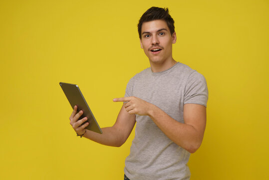 A surprised man in grey t-shirt against bright yellow backdrop holds tablet in left hand, pointing to screen with right hand, looking intrigued