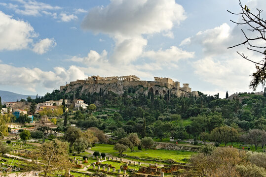 Panoramic view of the ancient Acropolis and the Parthenon temple sitting atop the hill, overlooking the ruins of the Ancient Agora and the Church of the Holy Apostles under a cloudy blue sky