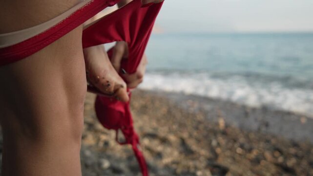 Beach change bikini. Woman pulling off her red swimsuit bottoms while standing on a rocky shoreline near the ocean.