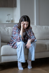 Obraz premium Anxious stressed young woman holding head with tense, lost in depressed thoughts. Worried female sits alone on sofa, touching temple with hand. Mental fatigue, loneliness, inner conflict, frustration.