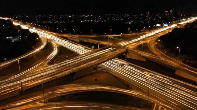 An aerial long exposure captures the glowing light trails of vehicles crossing a complex highway interchange at night.