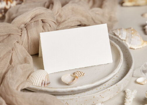 Mockup of folded card on plate with seashells, coral and beige fabric, coastal wedding stationery