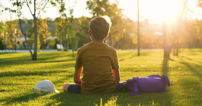 Teen enjoys calm pause on grass at sunset. Teenager sits on grass during golden hour. Backpack lies near on lawn with cap close by. Scene shows safety, rest, reflection. Choolboy in summer park.