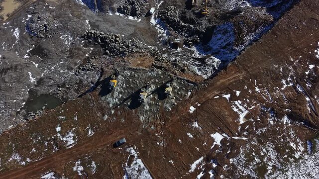 Heavy machinery is operating in a granite quarry. Workers are involved in extracting granite from the ground. The site shows disturbance and activity indicative of mining.
