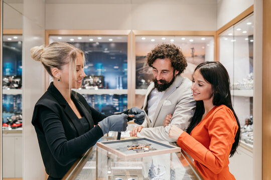 A professional sales assistant in a luxury jewelry store wearing gloves shows a selection of premium watches to a young couple during a high-end retail shopping experience.