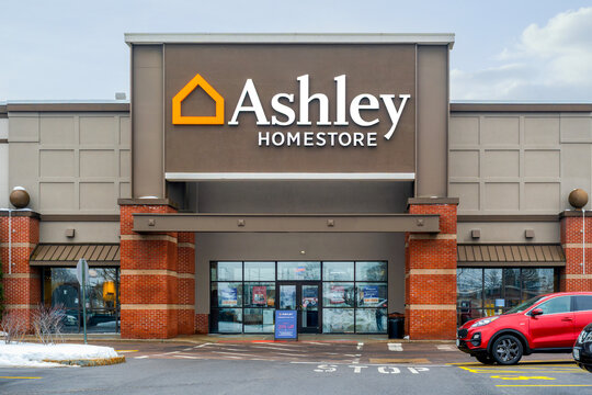 Syracuse, NY - Feb 17, 2026: Ashley HomeStore exterior with orange house‑icon logo, brick‑and‑gray facade, and winter surroundings including wet pavement and snow patches