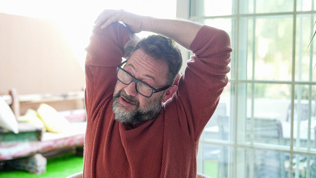Active mature bearded man exercising inside home with stretching exercises for shoulders and arms in a modern living room. Healthy  lifestyle, strength training, and wellness concept