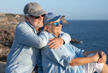 Fototapeta premium Happy senior couple hugging and smiling while sitting on a stone wall by the sea. Enjoying retirement, love, and companionship during a sunny coastal vacation