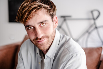 Obraz premium Happy young adult man with beard sitting on a sofa at home looking at camera in a relaxed modern living room setting, bright light from the window